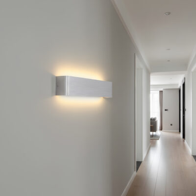 Lampe murale aluminium brossé gris allumée et diffusant une lumière jaune au-dessus et au-dessous. Elle est fixée dans un couloir.