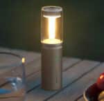 Une lampe de poche en forme de tube gris avec une lumière jaune posée sur une table en bois en extérieur, à gauche un verre d'eau.