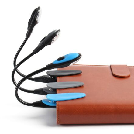 Un agenda en cuir marron posé sur un fond blanc. Trois lampes de lecture accrochées avec leur pince à l'agenda. Une lampe bleue, une grise et une noire. Sur fond blanc.