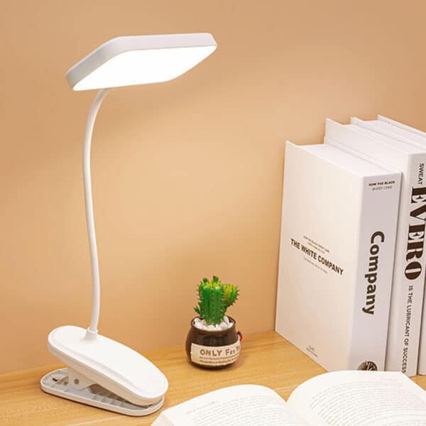 Une lampe de table à pince blanche posée sur un bureau en bois. Une petite plante à côté, deux livres et un livre ouvert. Sur fond beige.