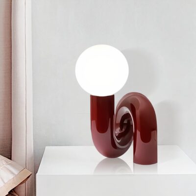 Une lampe de chevet design, le pied en forme de tube en résine rouge, l'abat-jour en forme de boule blanche. posée sur une table de chevet blanche laquée, sur fond blanc.