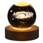 Une lampe boule de cristal posé sur un socle en bois rond, dans la boule, il y a la représentation d'une galaxie éclairée par le socle. Sur fond blanc.