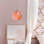 Dans une chambre près d'un lit, un luminaire suspendu en forme de fleur rose et dorée, se trouve devant un miroir