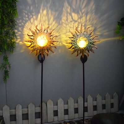 Deux lampes solaires en forme de soleil design planté dans le sol devant la façade d'une maison. Derrière elle, il y a une petite balustrade en bois et des buissons de chaque côté.