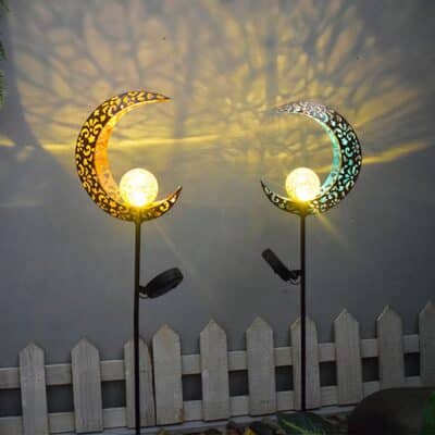 Deux lampes solaires en forme de lune design planté dans le sol devant la façade d'une maison. Derrière elle, il y a une petite balustrade en bois et des buissons de chaque côté.