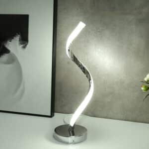 Lampe tige en spirale, en métal et en bande LED sur toute la tige. Son socle rond est en métal. Posée sur un meuble blanc, on voit un mur gris en fond et un bout de cadre sur la gauche.