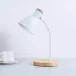 Lampe de bureau blanche en fer, avec le socle rond en bois et une petite partie derrière l'abat-jour en bois. Posée sur une table grise avec une tasse et une sous tasse sur la gauche.