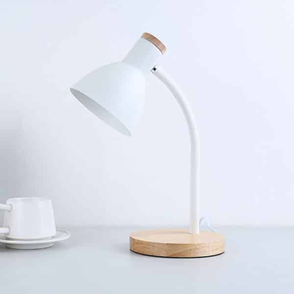 Lampe de bureau blanche en fer, avec le socle rond en bois et une petite partie derrière l'abat-jour en bois. Posée sur une table grise avec une tasse et une sous tasse sur la gauche.