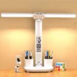 Lampe de bureau blanche, posée sur une table en bois, avec une horloge intégré dans la tige. Le socle dispose de deux portes craillons intégrés, avec des crayons. La lumière est allumée et en forme de tige pliable. On voit un mur jaune en fond. Sur la gauche un petit robot chien et sur la droite un cubicule.