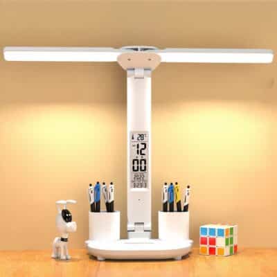 Lampe de bureau blanche, posée sur une table en bois, avec une horloge intégré dans la tige. Le socle dispose de deux portes craillons intégrés, avec des crayons. La lumière est allumée et en forme de tige pliable. On voit un mur jaune en fond. Sur la gauche un petit robot chien et sur la droite un cubicule.