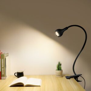Photo d'une table en bois sur laquelle est posé un livre ouvert, avec une tasse, éclairé par une lampe pince accrochée sur le côté droit. On voit un mur gris dans le fond et un petit pot avec une plante sur la droite.
