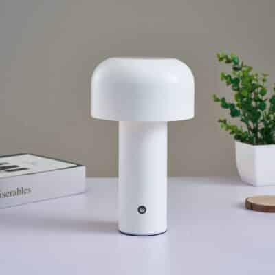Lampe blanche simple en forme de champignon avec un long pied. Posée sur une table blanche, avec un mur gris en fond, on voit à droite un petit pot de fleur et à gauche un livre.