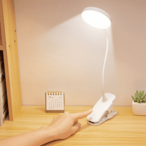 Lampe LED blanche à pince avec tige flexible et grand rond lumineux. Allumée et posée sur une table en bois, à sa droite un petit pot avec une plante verte, à sa gauche un calendrier. On voit un bras avec un doigt qui va appuyer sur l'interrupteur.
