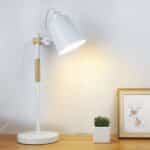 Lampe de bureau allumée, blanche avec un socle rond, un bras fixe avec une partie en bois, un abat-jour en cône blanc rotatif. Posée sur une table en bois qui éclaire un cadre posé contre le mur et un mini pot blanc avec une plante verte. On voit un mur beige dans le fond.