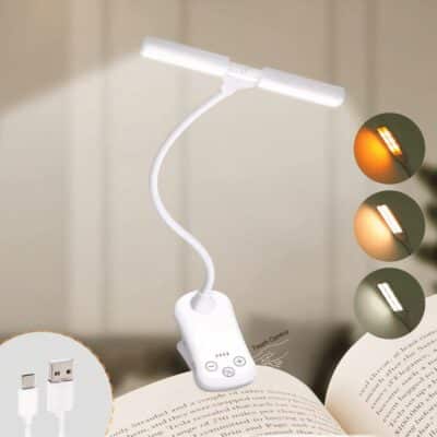 Lampe de lecture blanche à pince, accrochée à la page d'un livre. Sa tige est flexible. En bas à gauche un cercle avec les 2 embouts USB et sur le côté droit 3 cercle avec les différentes luminosités possible.