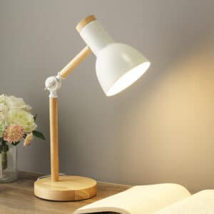 Lampe de bureau avec socle et pied en bois, l'abat-jour est en fer blanc. Elle est posée sur une table et éclaire un cahier ouvert. On voit un mur gris dans le fond et un bout de bouquet de fleur sur la droite.