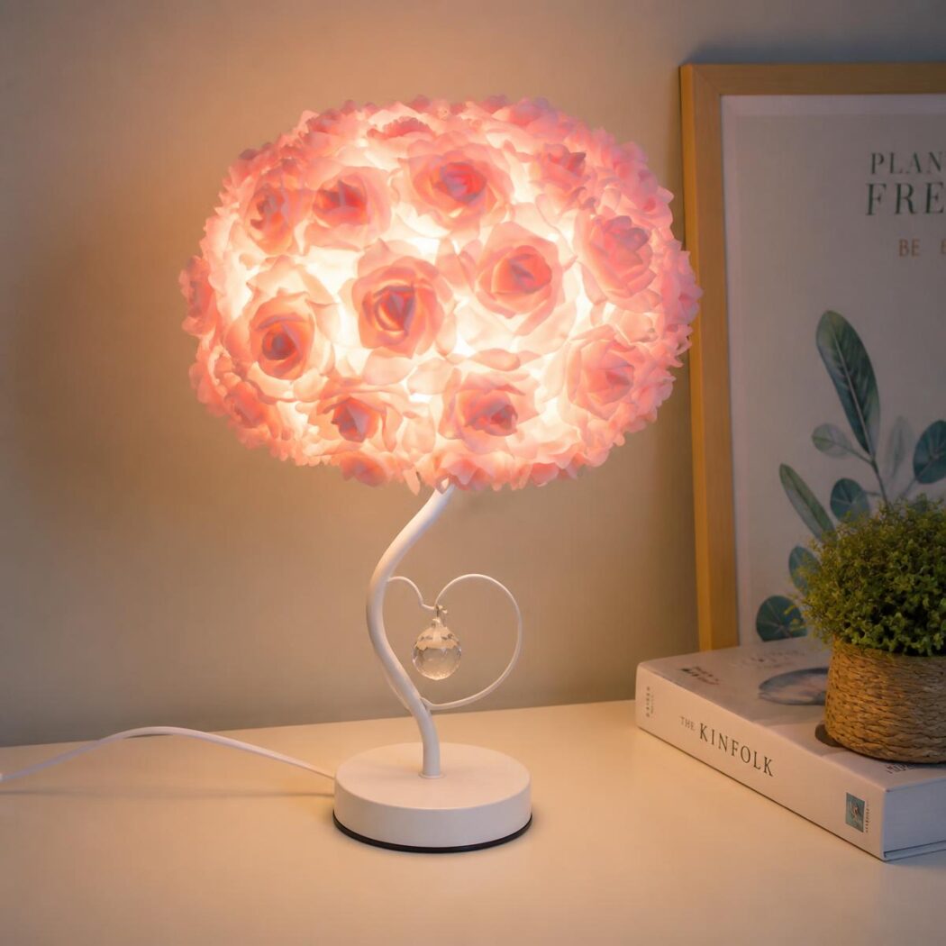 Une lampe fleur bouquet de roses rose posée sur une table de chevet, éclairant doucement un livre dans une chambre cosy.