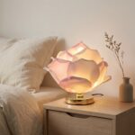 Une lampe fleur de lotus avec pétales en tissu rose et blanc, posée sur une table de chevet en bois dans une chambre douce et calme.