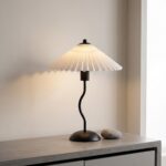 Une lampe ondulée rétro de table au charme chaleureux posée sur un meuble en bois, avec un abat-jour plissé blanc qui diffuse une lumière douce et un pied noir courbé.