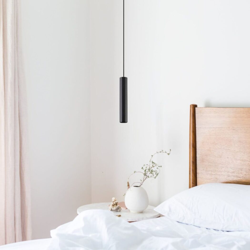 Une suspension tube verticale minimaliste pour îlot central noire, suspendue au-dessus d’un lit dans une chambre claire et épurée.