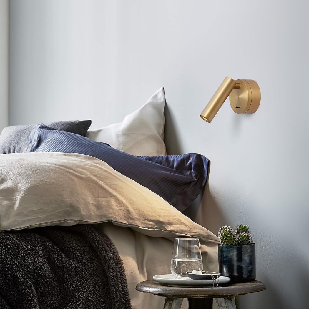 Une applique murale orientable en laiton design minimaliste fixée au mur, au-dessus d’un lit avec des coussins et une petite table près d’une plante en pot.