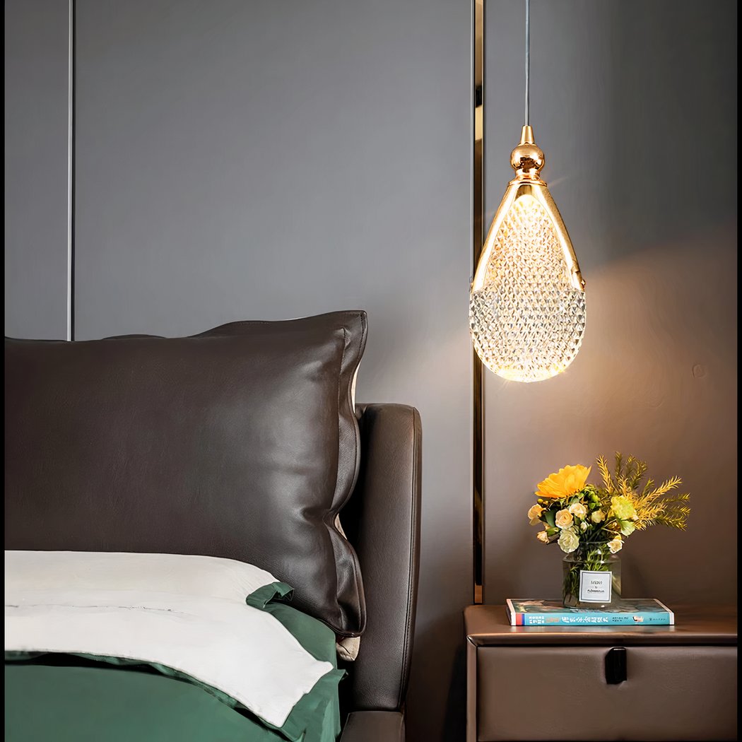 Une suspension goutte en cristal acrylique design contemporain brillante au-dessus d’une table de chevet, dans une chambre moderne aux tons gris et marron.
