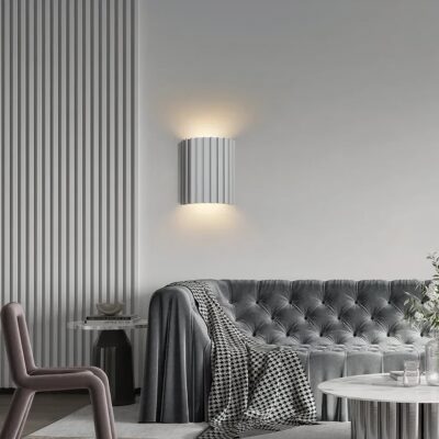 Une applique murale scandinave cylindrique striée à double éclairage fixée au mur, qui diffuse une lumière douce vers le haut et le bas dans un salon moderne gris.